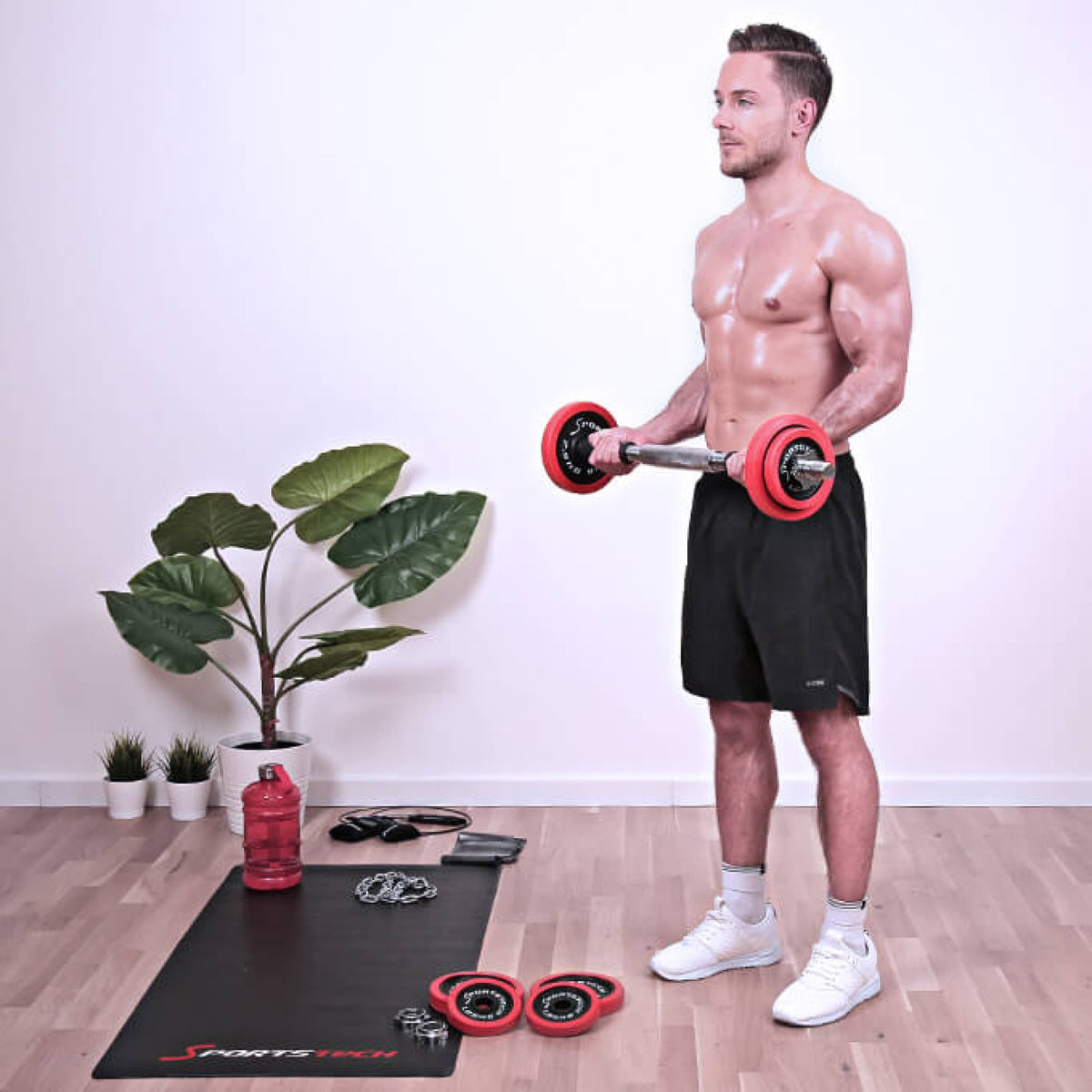 Stehender Mann macht Bizeps-Curls mit der AH300 Langhantelstange, rote Scheiben, im Wohnraum mit Matte