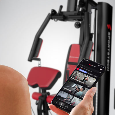 Smartphone mit Sportstech Live App in der Hand vor Polster und Gewichtsturm der HGX150 Kraftstation
