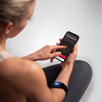 Frau mit sPulse Armband hält Smartphone mit App-Bildschirm „Verbinde dein Trainingsgerät!“, Sensor leuchtet blau