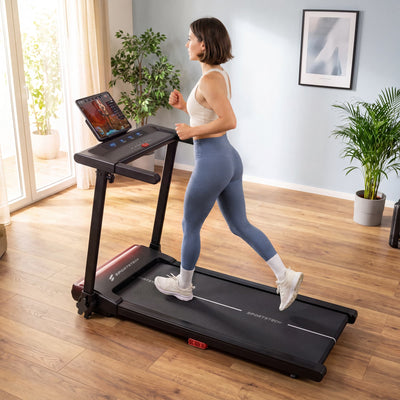 Frau in Sportkleidung joggt im Wohnzimmer auf dem sProRun Laufband, Tablet in der Konsolenhalterung