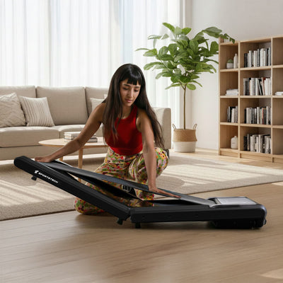 Frau klappt das sWalk Fold Walking Pad im Wohnzimmer in der Mitte zusammen