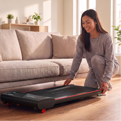 Frau schiebt das flache Walkmate Walking Pad unter ein Sofa im Wohnzimmer