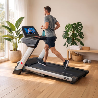 Mann joggt am F75 Profi-Laufband im Wohnzimmer mit Fenster, Pflanzen, Holzbank und Hanteln