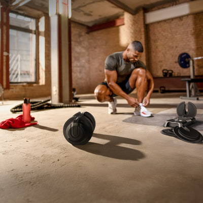 Ironmate Hantel liegt auf dem Studioboden, dahinter schnürt ein Sportler seine Schuhe