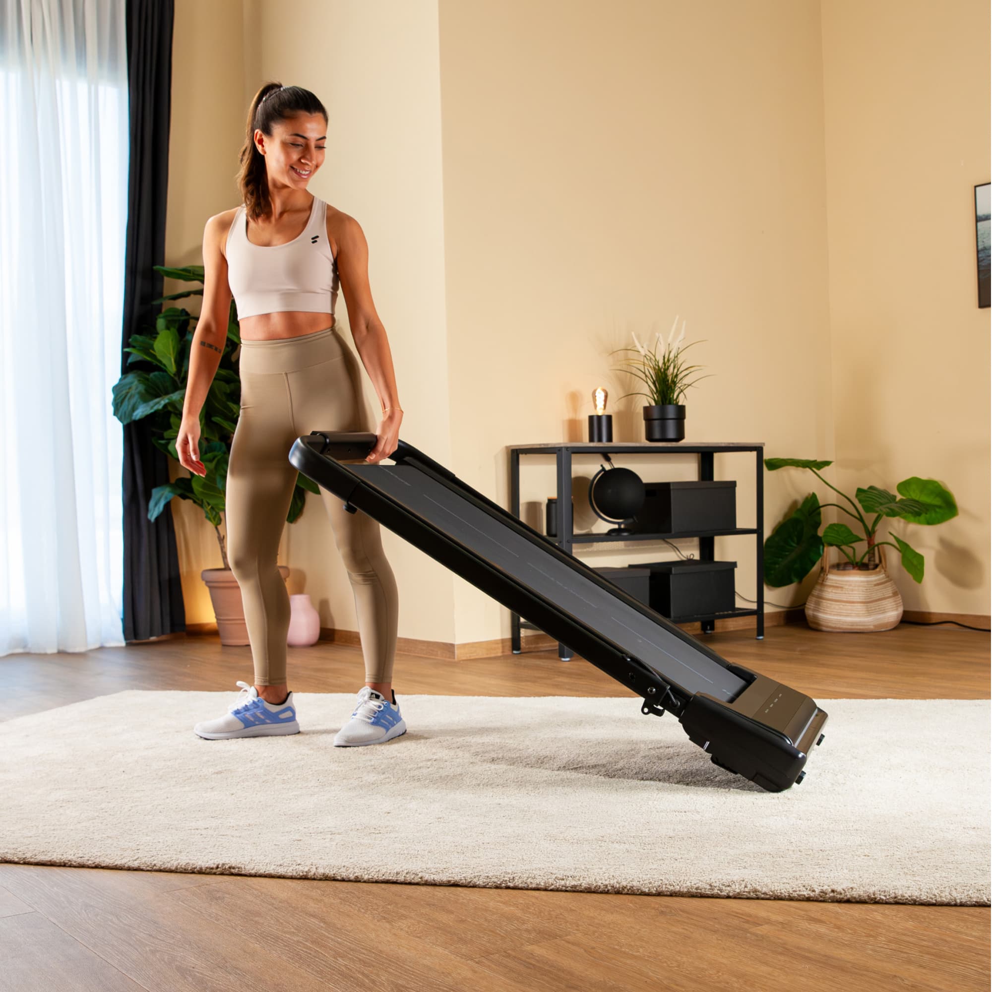 Frau klappt das sWalk Lite Plus Laufband am Haltebügel hoch, das schräg auf dem Teppich steht