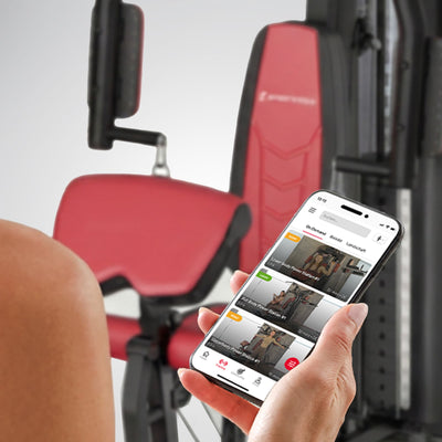 Hand hält Smartphone mit Trainings-App vor der roten Sitzpolsterung der HGX150 Kraftstation