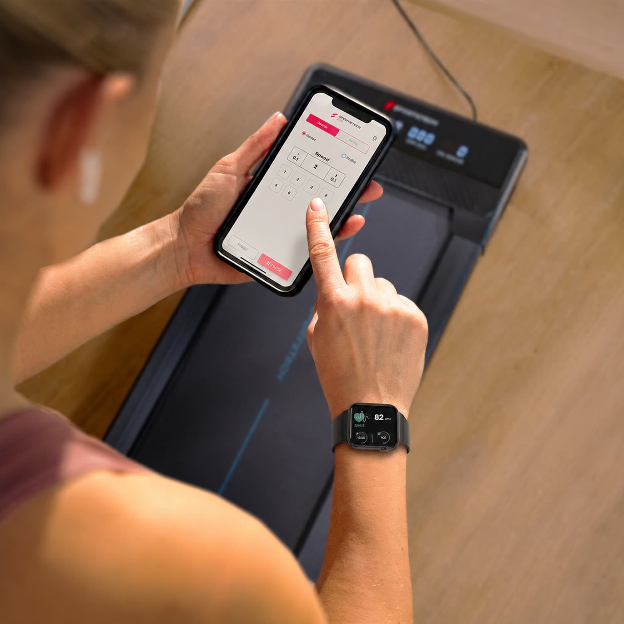Frau steuert das sPad500 Walking Pad über die Smartphone-App, am Handgelenk eine Smartwatch mit Puls
