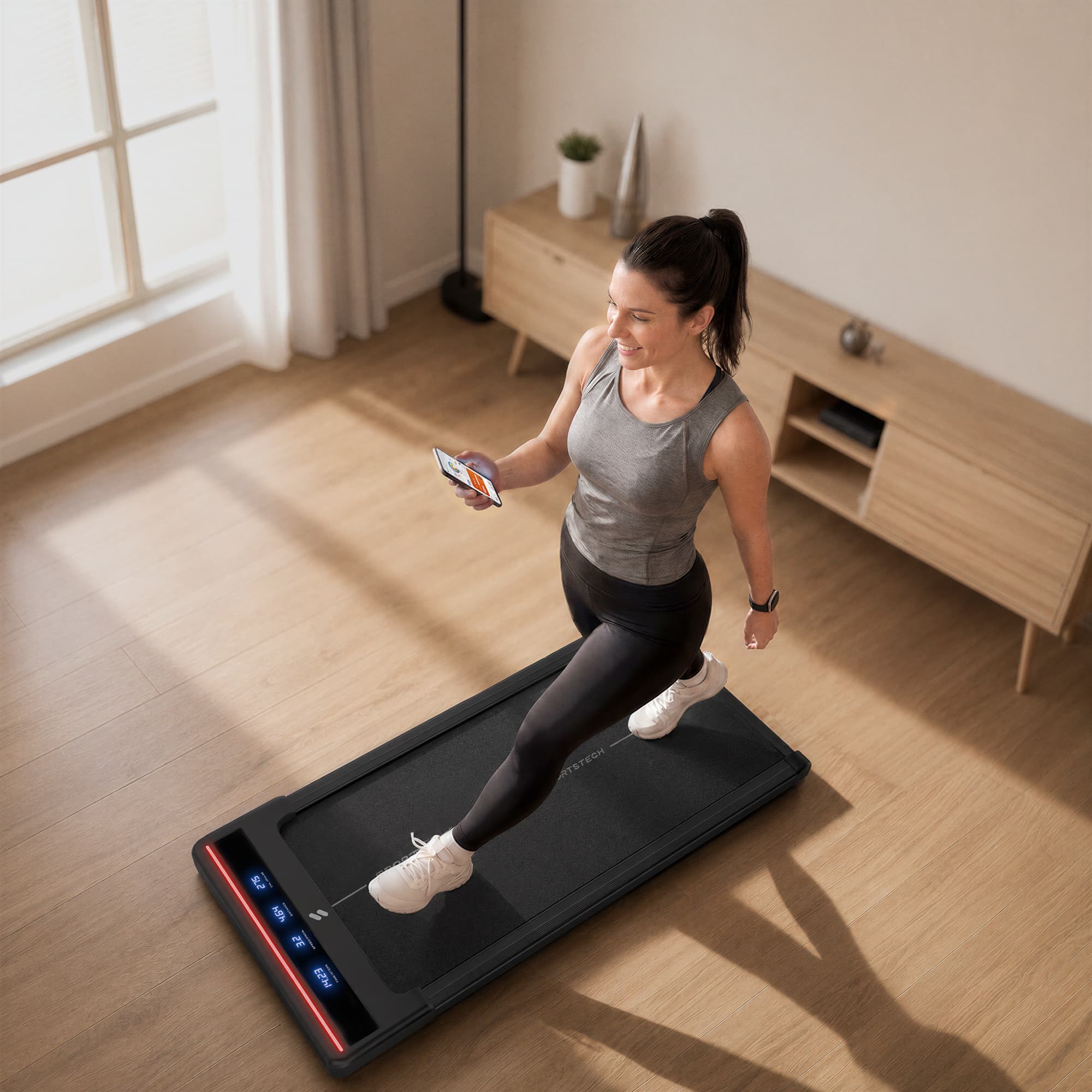 Frau mit Smartphone geht auf dem sWalk Lite Walking Pad mit rot leuchtender LED-Leiste im Wohnzimmer