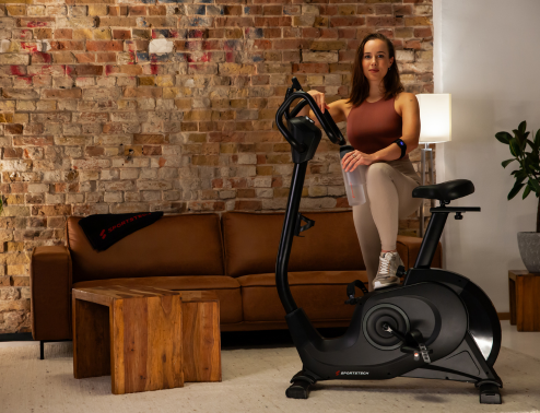 Eine Frau steht neben dem Sportstech Ergometer in ihrem Wohnzimmer
