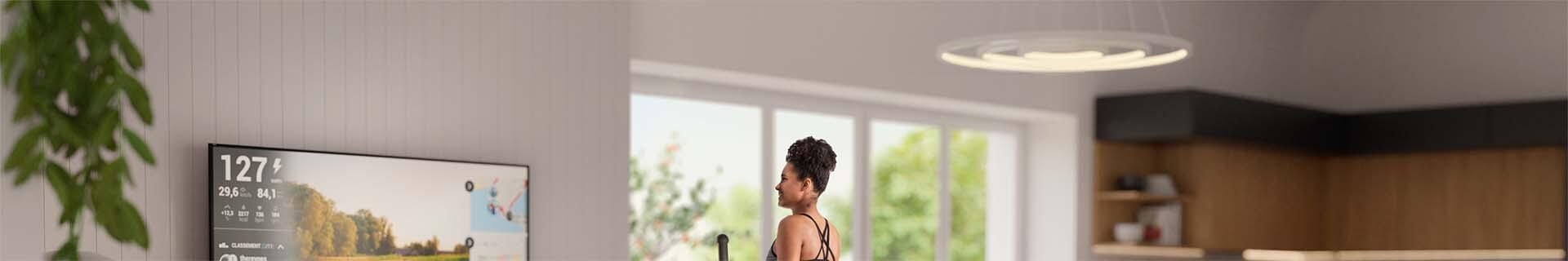 Crosstrainer CX625 von Sportstech Frau in Sportbekleidung trainiert auf dem CX625 Crosstrainer im Wohnzimmer und verwendet die Fitness App auf dem TV